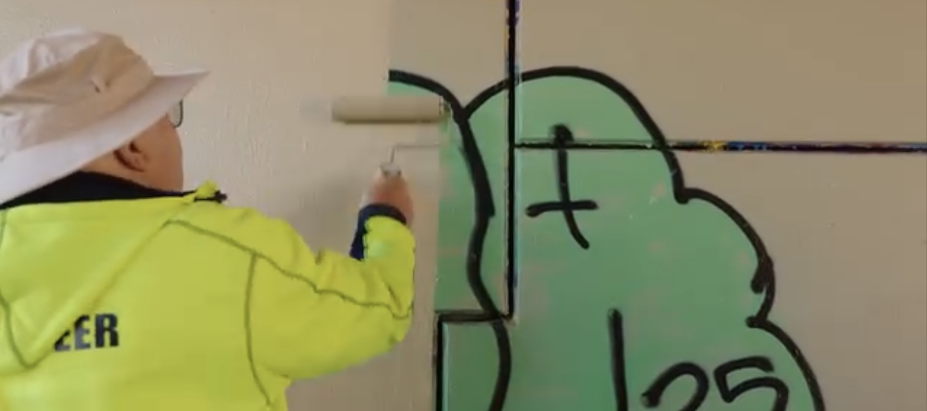 Man using beige paint on a roller to apply to graffitied wall.