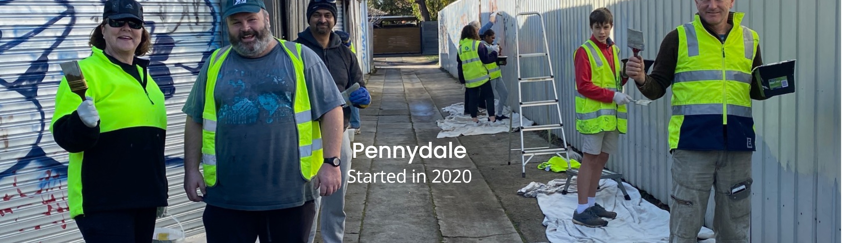 Group of neighbourhood volunteers in casual clothing and high viz vests painting over graffiti in a lane way. All smiling and happy.
